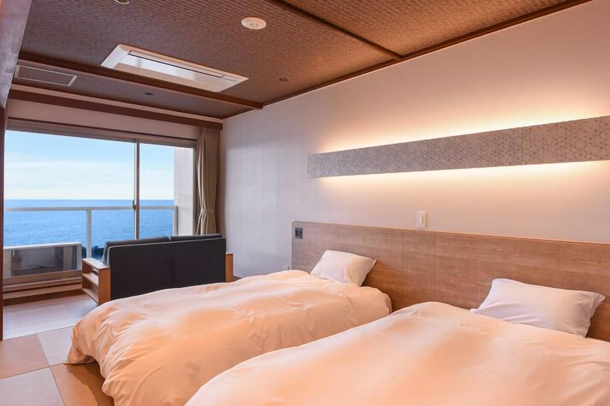 A hotel room with two beds, a desk, and a large window offering a view of the sea.