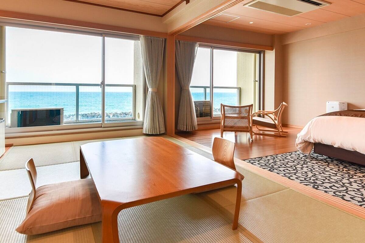 A hotel room with a bed, a dining table, chairs, a view of the sea, and large windows.