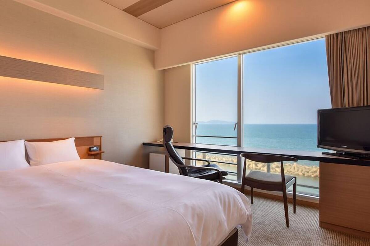 A hotel room with a bed, a desk with a chair, a TV, and a large window with a view of the sea.