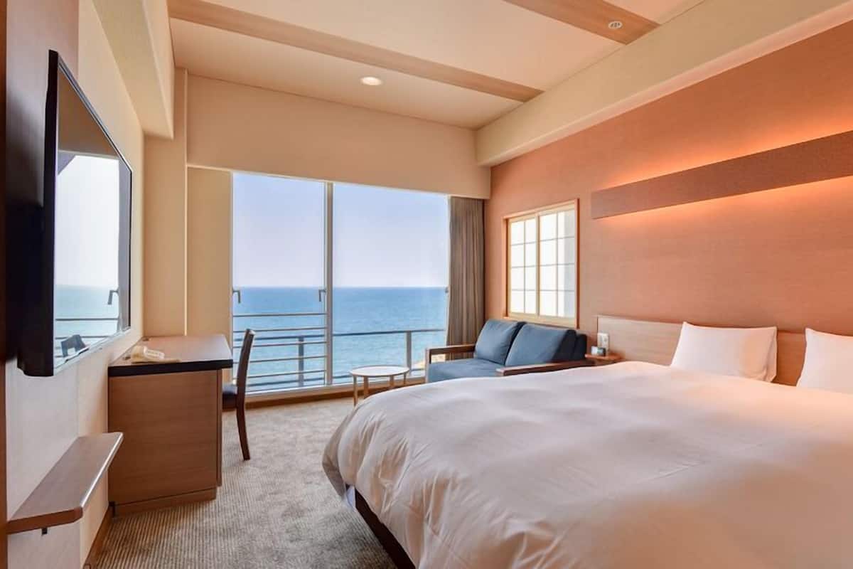 A hotel room with a large bed, a desk, a chair, a TV, and a balcony with a view of the sea.
