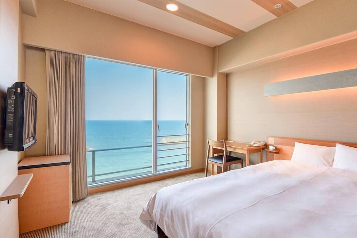 A hotel room with a bed, a desk, a chair, a TV, and a large window with a view of the sea.