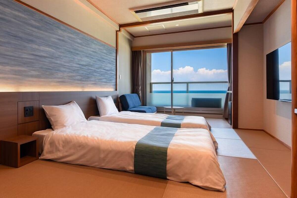 A modern hotel room with two beds, a large window with a view of the sea, and a flat-screen TV.