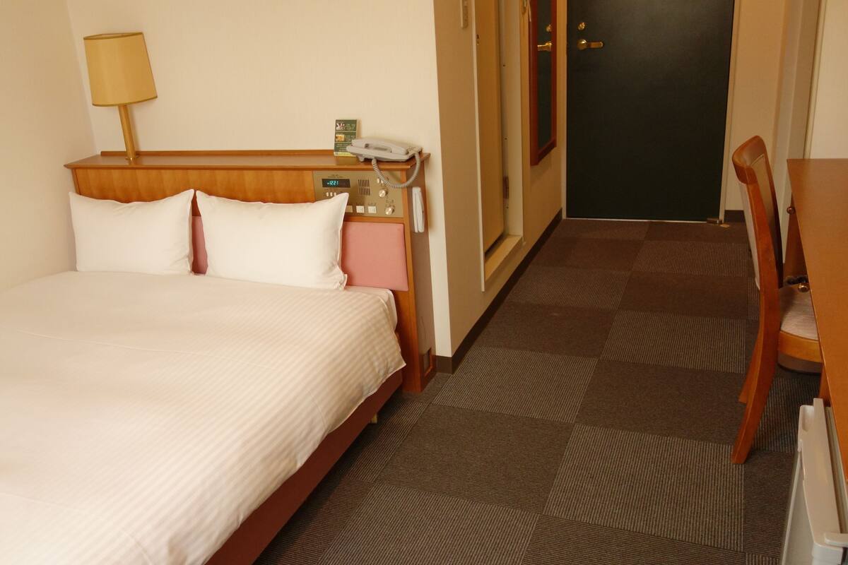 A hotel room with a bed, a bedside table with a lamp, a phone, and a door.
