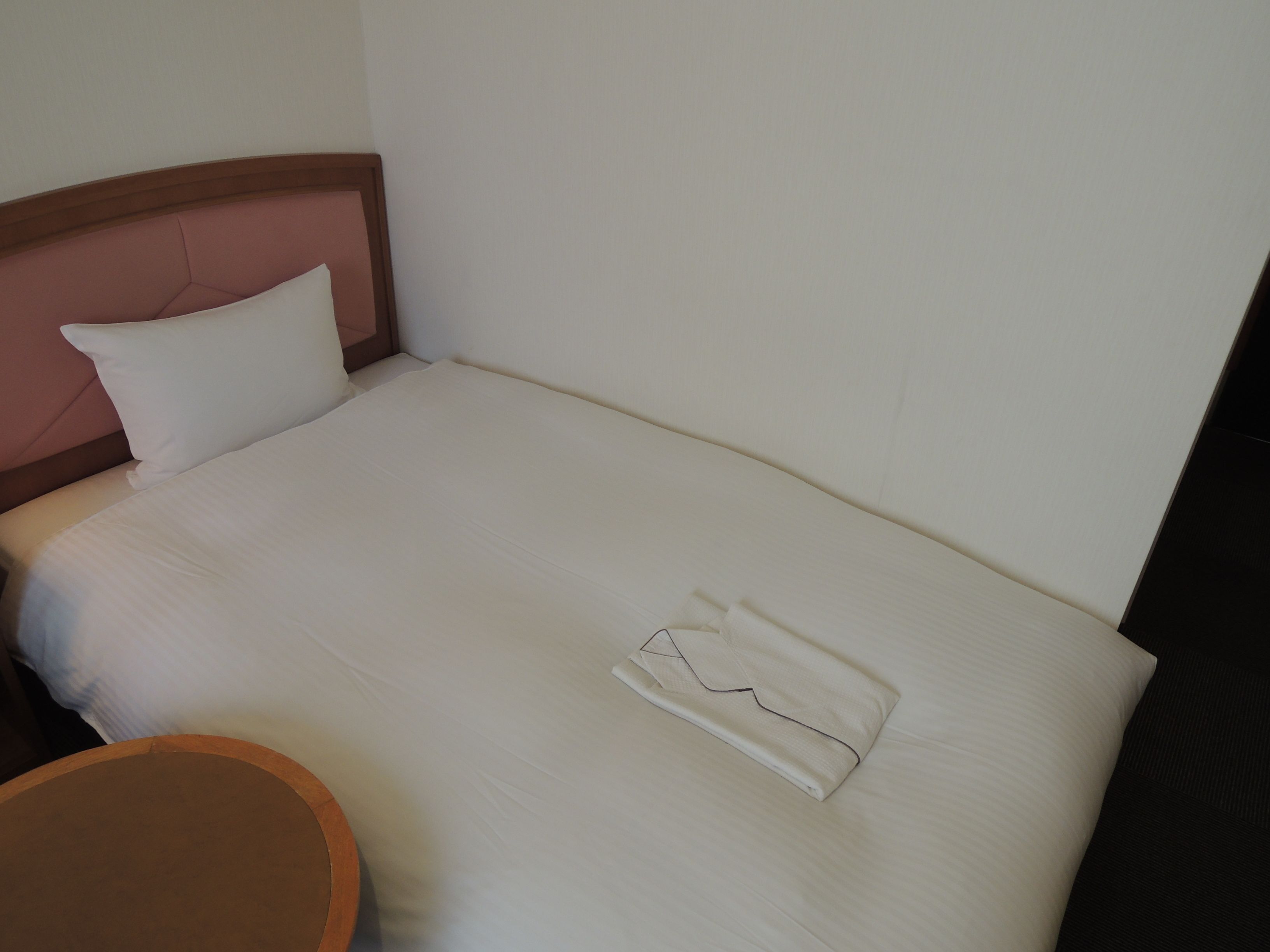 Una cama bien tendida con una toalla doblada y una almohada, una mesa redonda de madera y una pared lisa.