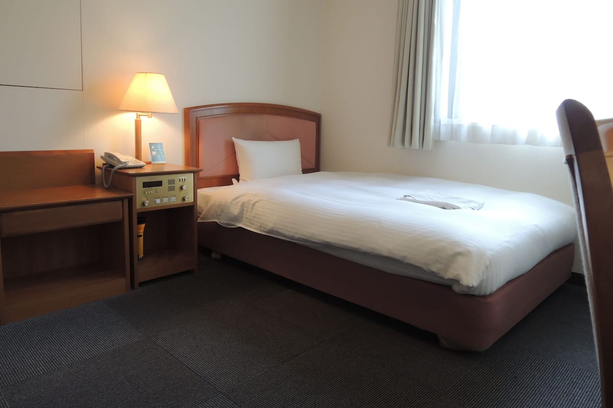 A hotel room with a bed, a desk with a telephone, a lamp, and a window with curtains.
