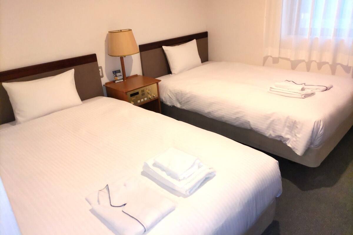 Two single beds with white linens, a bedside table with a lamp, and a window with curtains.