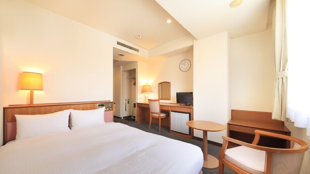 Habitación de hotel con una cama grande, un escritorio con silla, una mesita, televisión y un reloj en la pared.