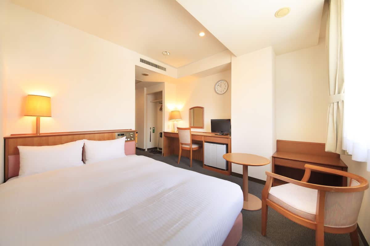 A hotel room with a large bed, a desk with a chair, a small table, a television, and a clock on the wall.