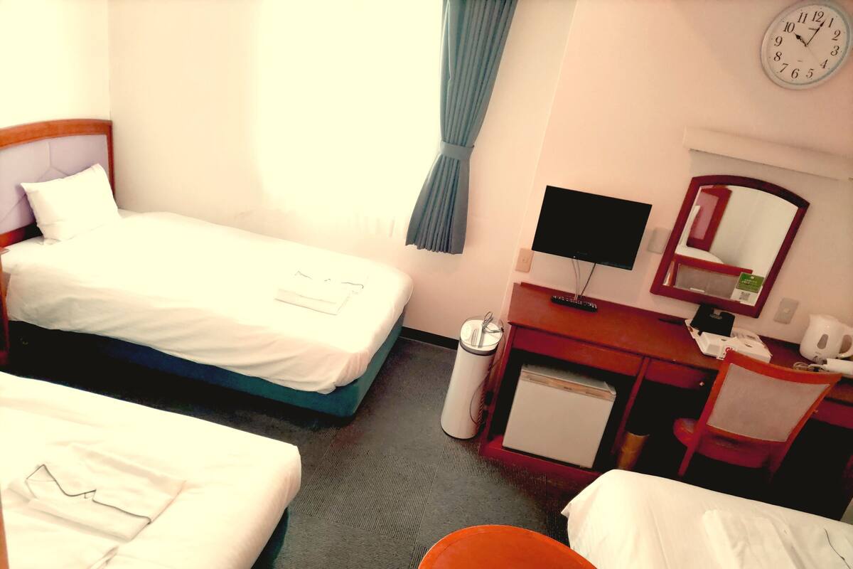 A hotel room with two beds, a desk with a computer, a small fridge, a trash bin, and a clock on the wall.
