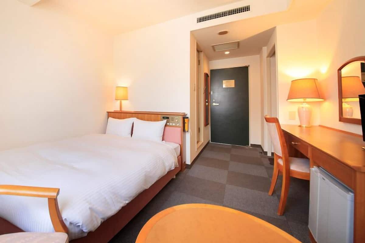 A hotel room with a bed, a desk, a chair, a lamp, and a door.