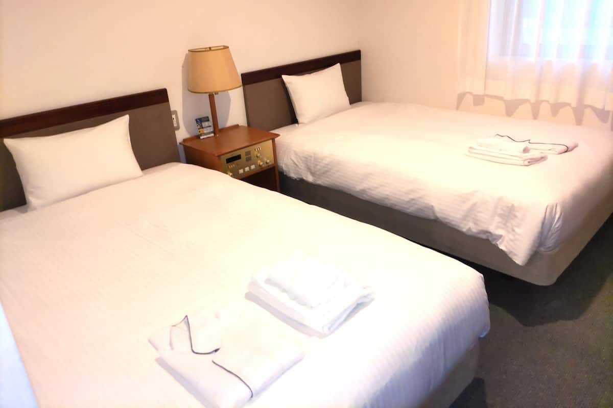 A hotel room with two beds, each with white linens and pillows, a nightstand with a lamp, and a window with curtains.