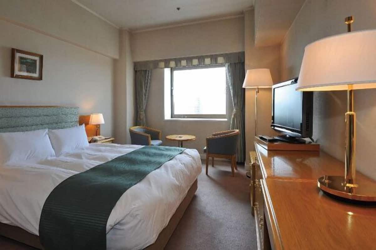 Une chambre d’hôtel avec un grand lit, un bureau avec une lampe, une télévision et une fenêtre avec des rideaux.