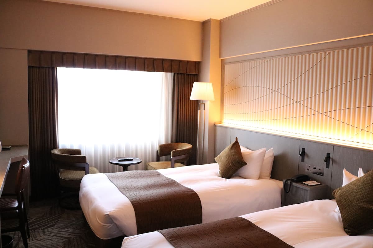 Une chambre d’hôtel avec deux lits, un bureau, une chaise, une lampe et une fenêtre avec des rideaux.
