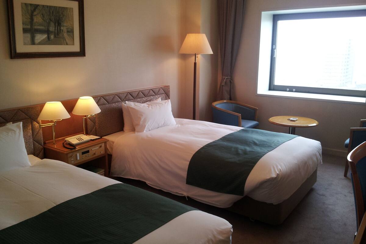 Une chambre d’hôtel avec deux lits, une table de chevet avec des lampes, une chaise, une petite table et une fenêtre donnant sur un paysage agréable.