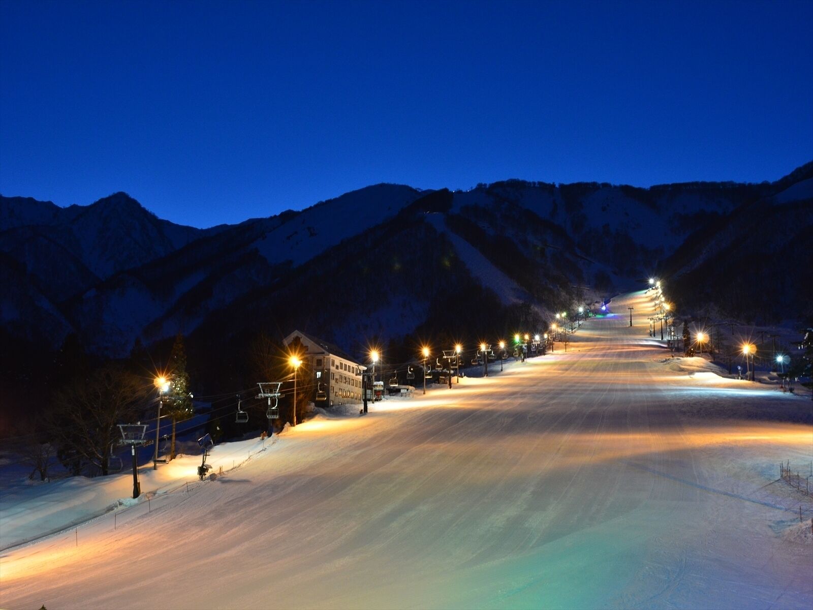 A snow-covered ski slope at dusk with illuminated buildings and a mountain backdrop.