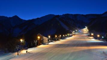 A snow-covered ski slope at dusk with illuminated buildings and a mountain backdrop.