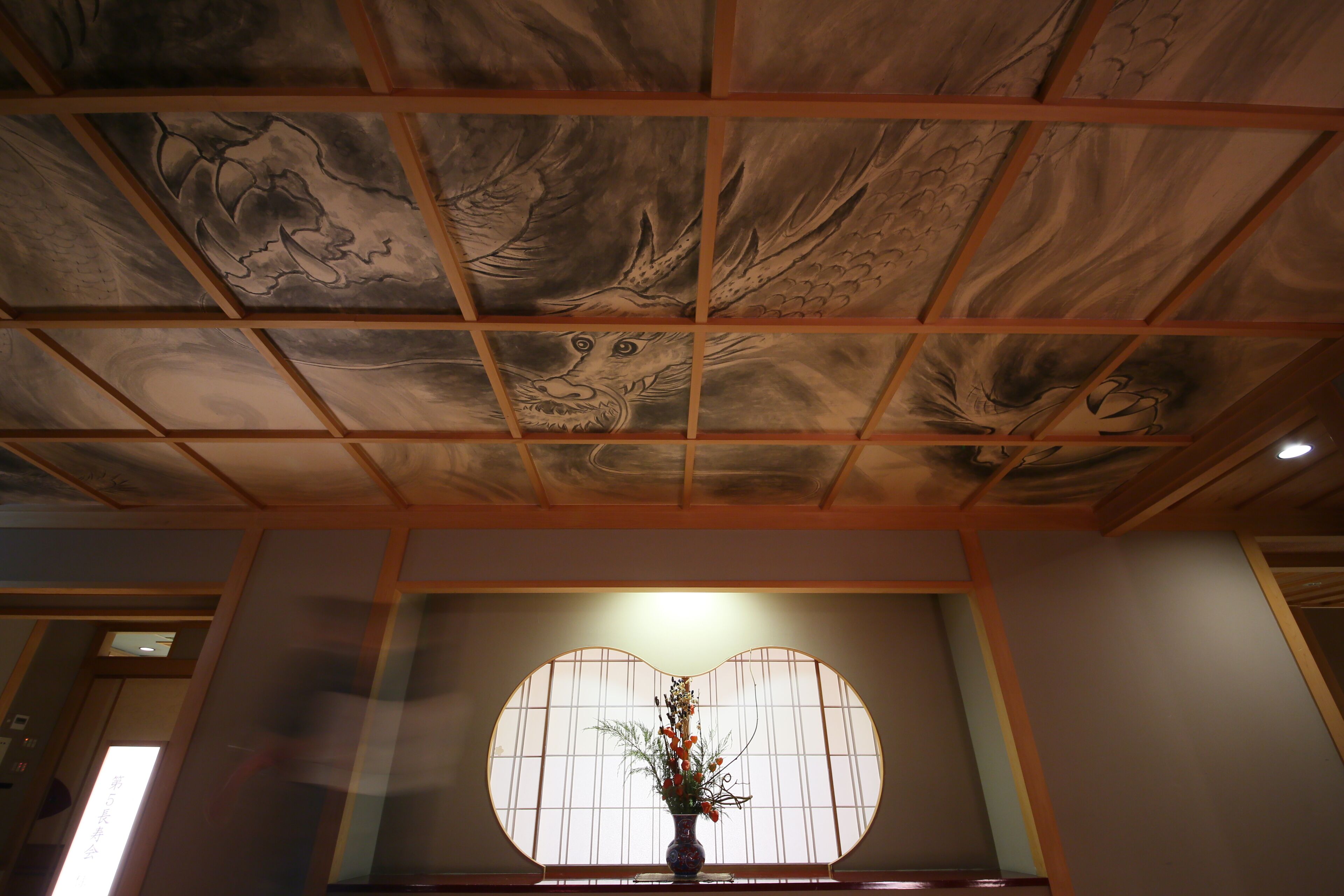 A room with a traditional ceiling design featuring dragon motifs, a shoji screen door, and a vase with flowers.