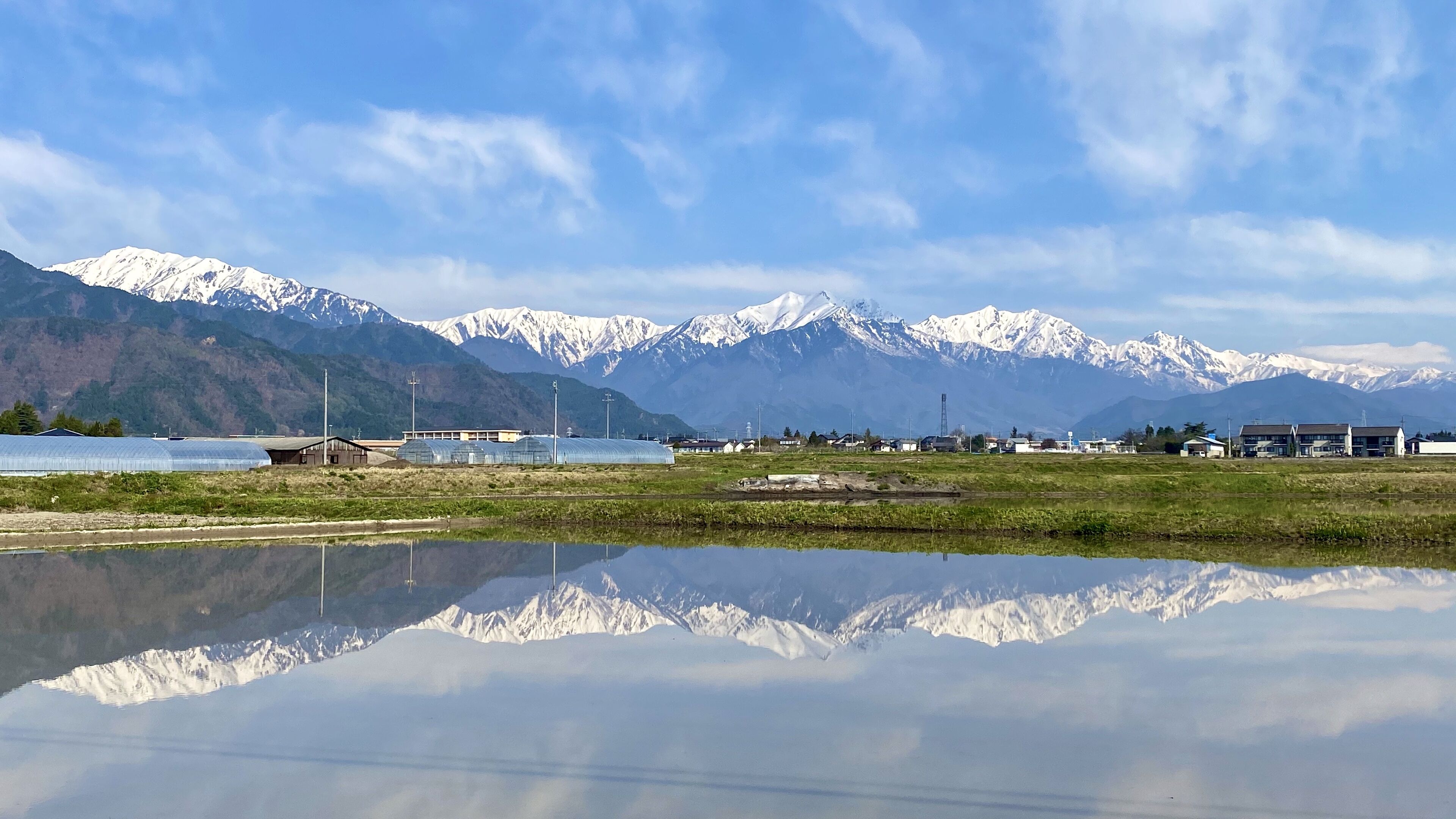 A mountainous landscape with a clear sky, a body of water reflecting the scenery, and several buildings in the foreground.