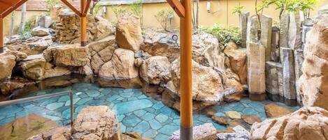 Ein Steingarten mit klarem Wasserspiel, Holzbalken und einer gelben Wand im Hintergrund.