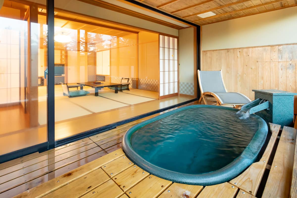 A traditional Japanese onsen with a wooden deck, a stone hot spring, and a view of a tatami room with furniture.