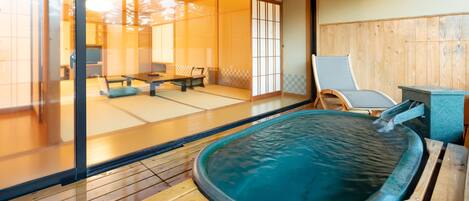 A traditional Japanese onsen with a wooden deck, a stone hot spring, and a view of a tatami room with furniture.
