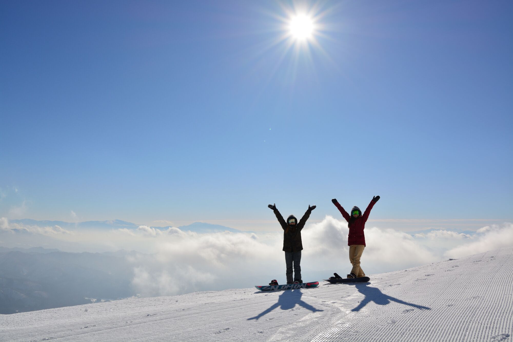 Deux personnes se tiennent sur une montagne enneigée, les bras levés, face à un ciel bleu dégagé et au soleil.