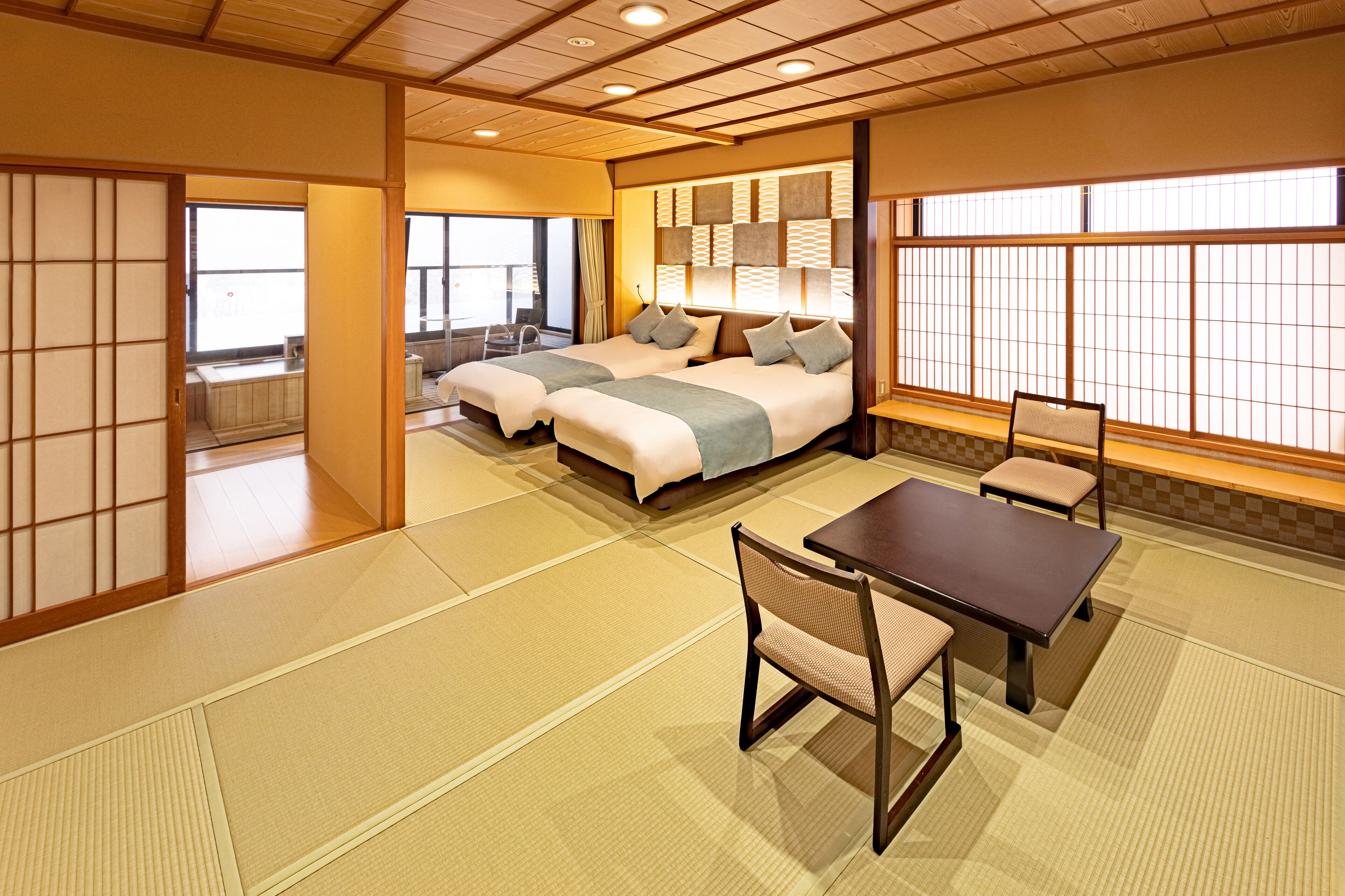A traditional Japanese-style room with tatami flooring, a low table with a chair, and a bed with pillows.