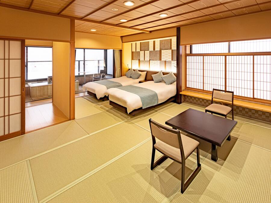 A traditional Japanese-style room with tatami flooring, a low table with a chair, and a bed with pillows.