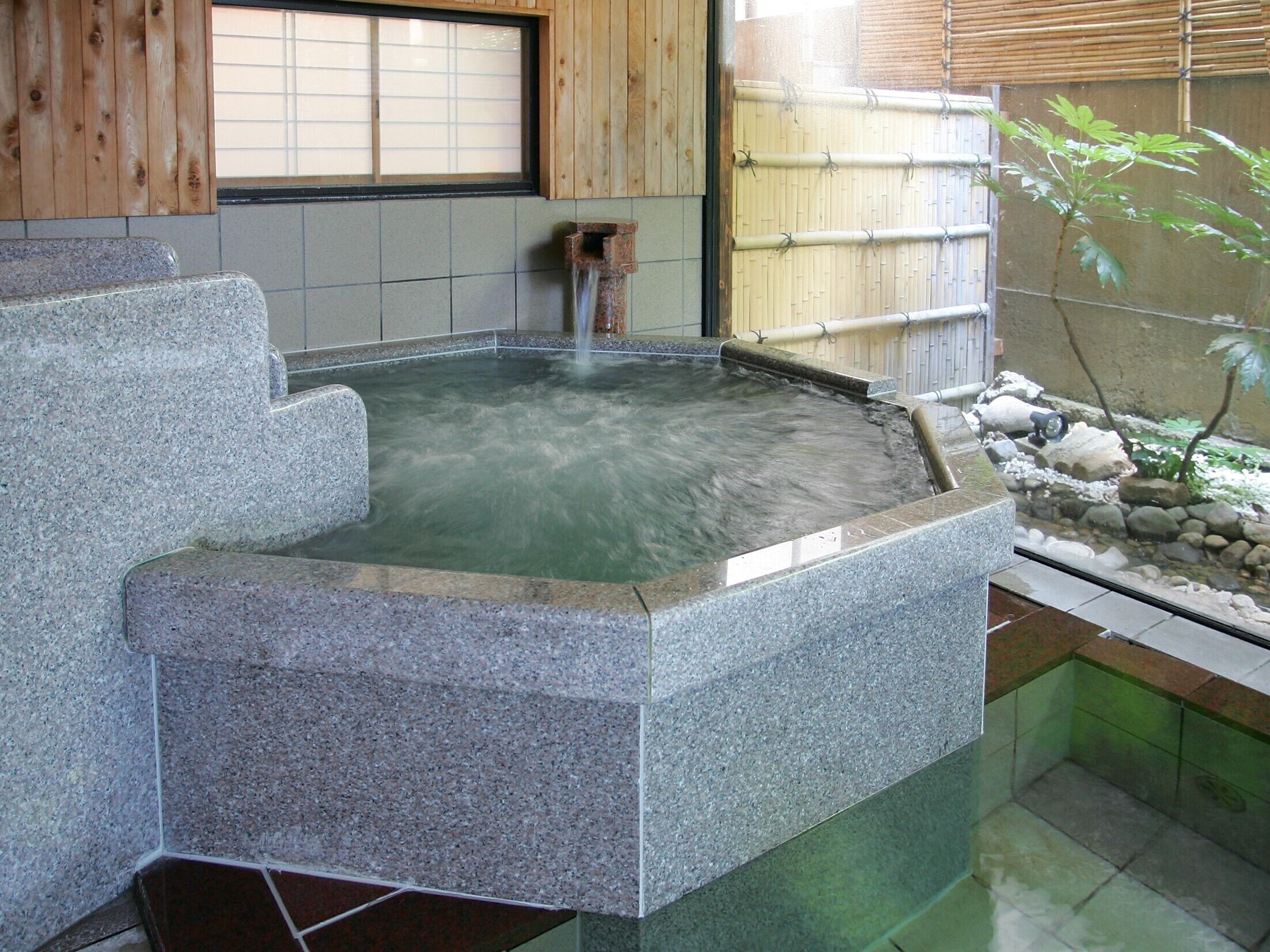 A stone hot spring bath with a wooden bench, a bamboo fence, and a small waterfall feature.