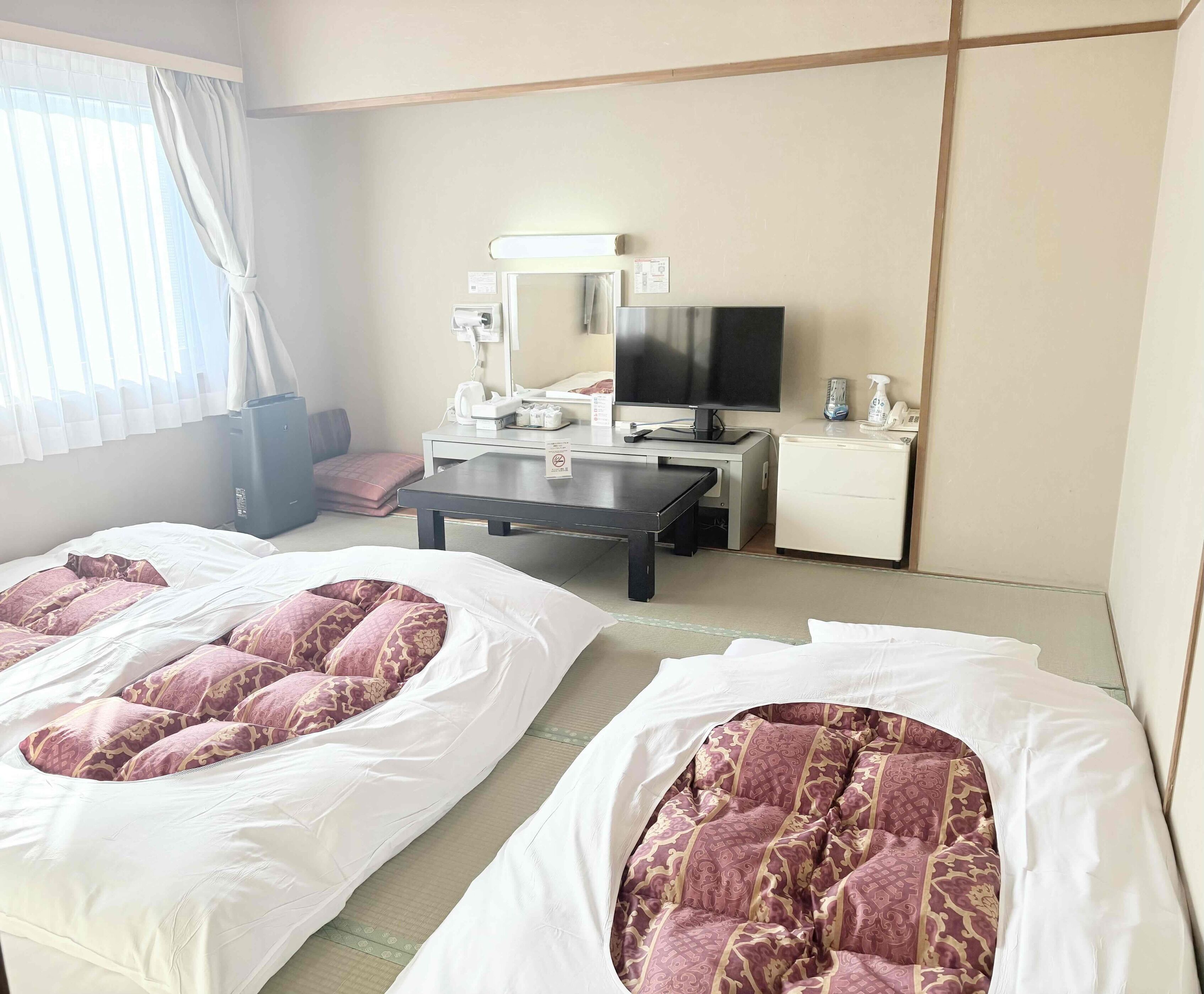 Quarto de hotel com duas camas, uma escrivaninha, televisão, pequeno frigorífico e janela com cortinas.