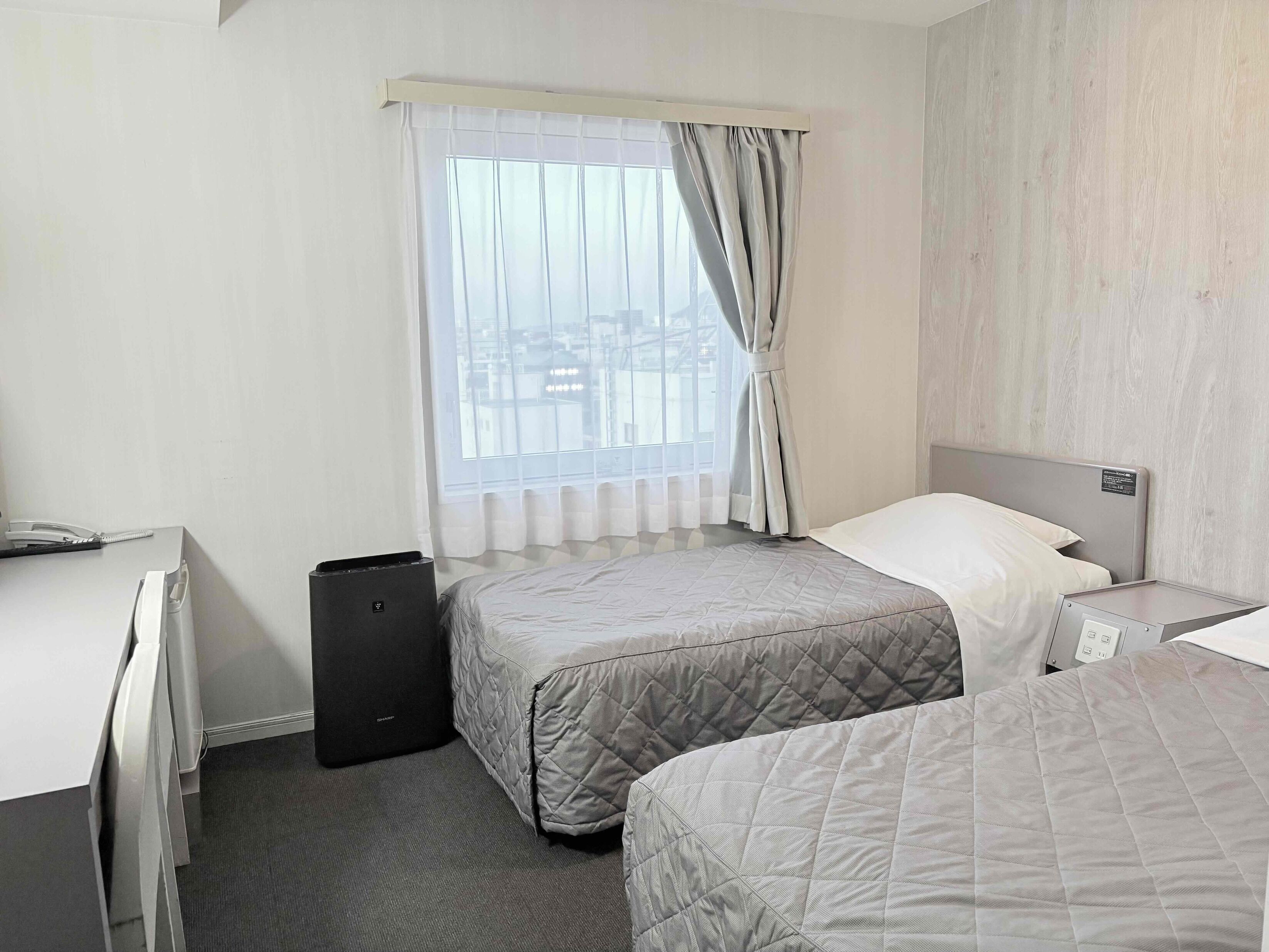 Quarto de hotel com duas camas, uma escrivaninha, uma cadeira, uma mala preta e uma janela com cortinas.
