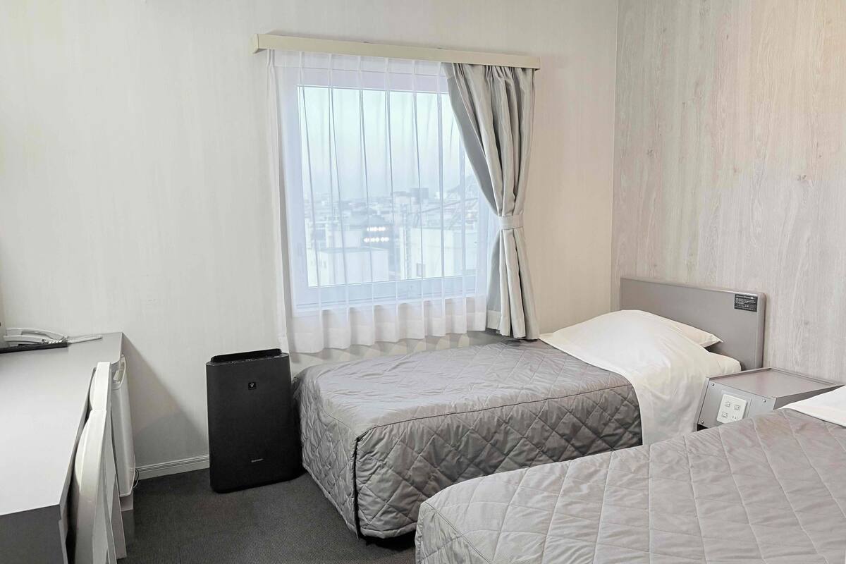 Quarto de hotel com duas camas, uma escrivaninha, uma cadeira, uma mala preta e uma janela com cortinas.