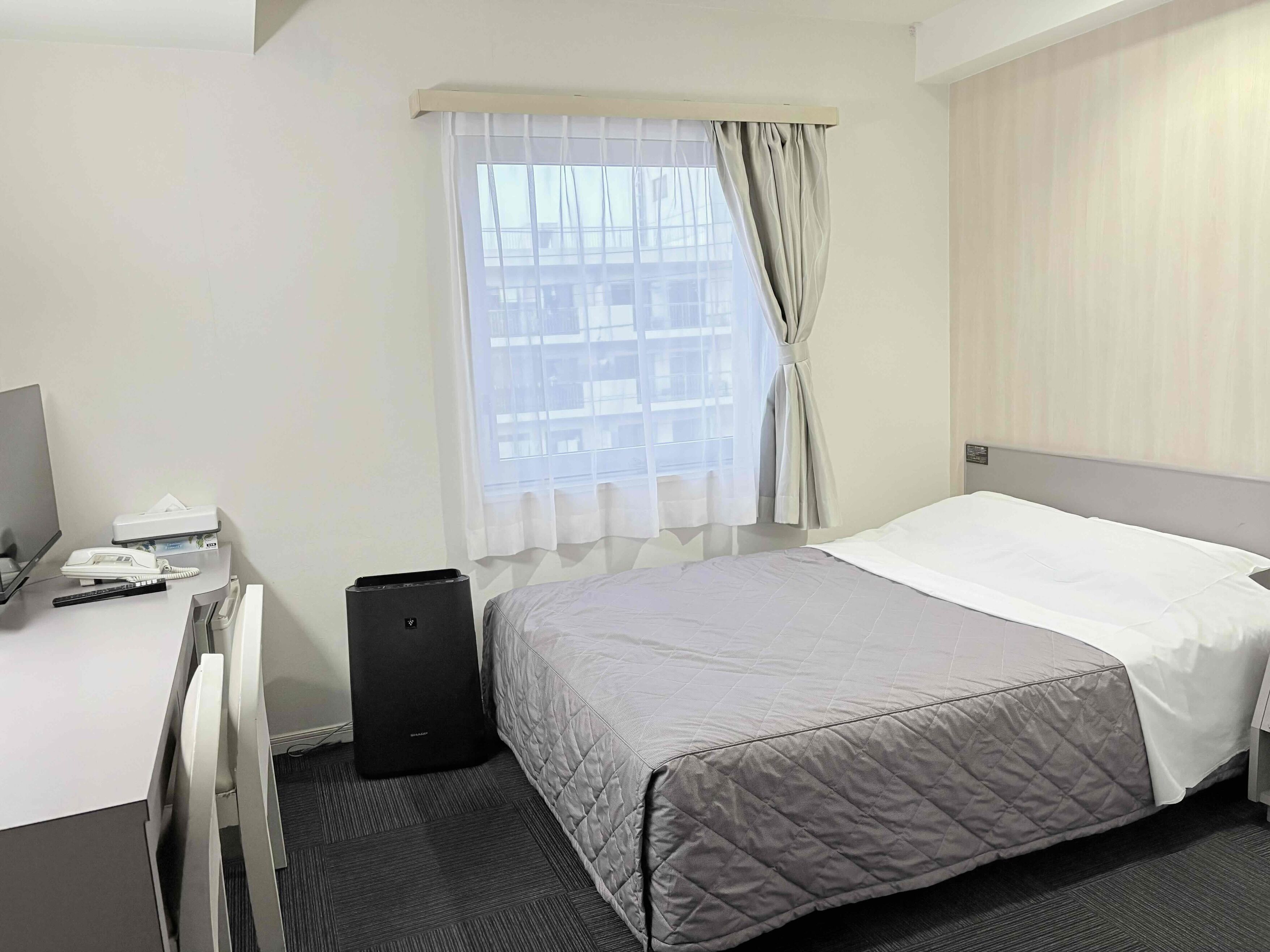 Quarto de hotel com cama, escrivaninha, telefone, alto-falante e janela com cortinas.