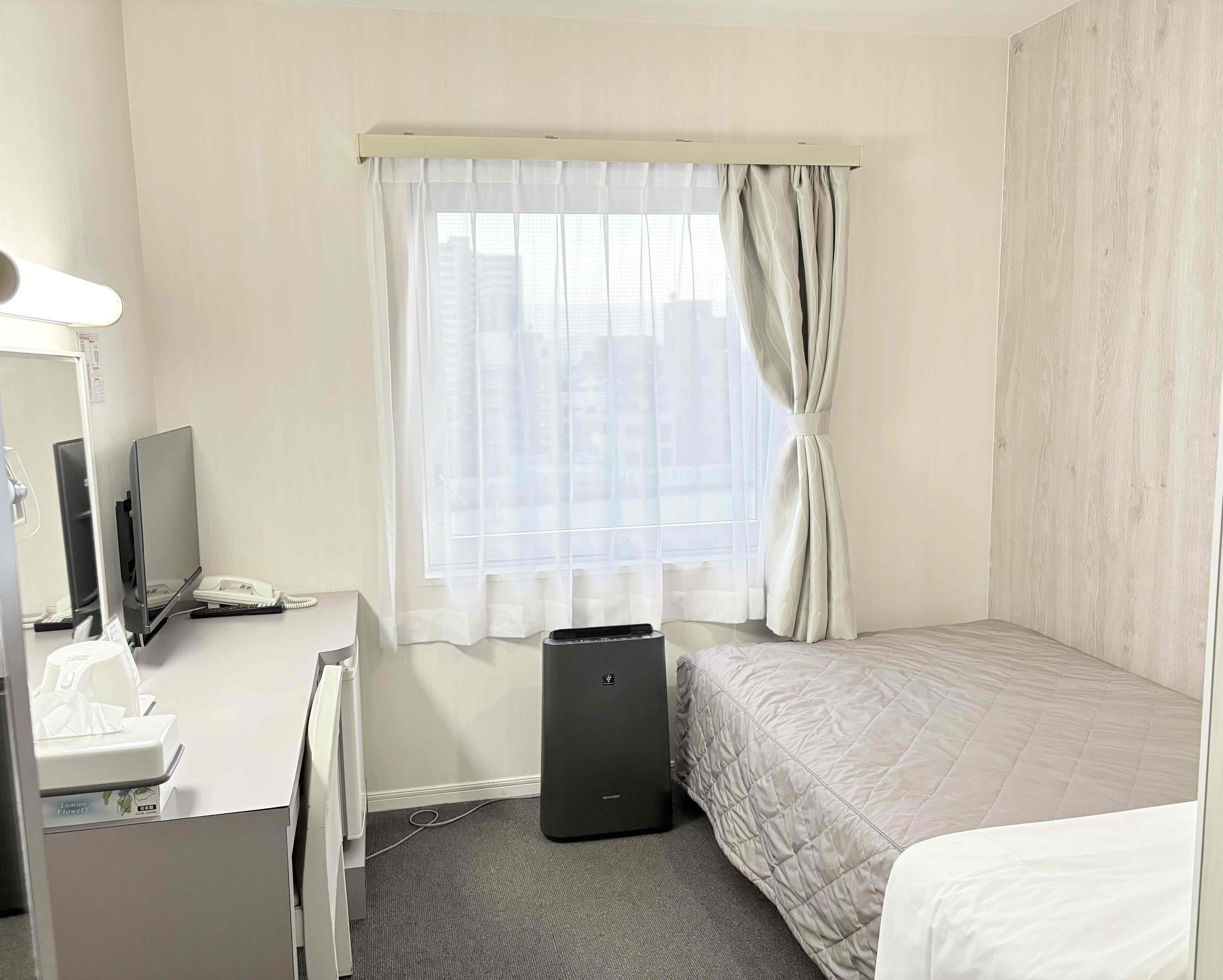 Quarto de hotel com uma cama, uma escrivaninha com computador, um telefone e uma pequena lixeira.