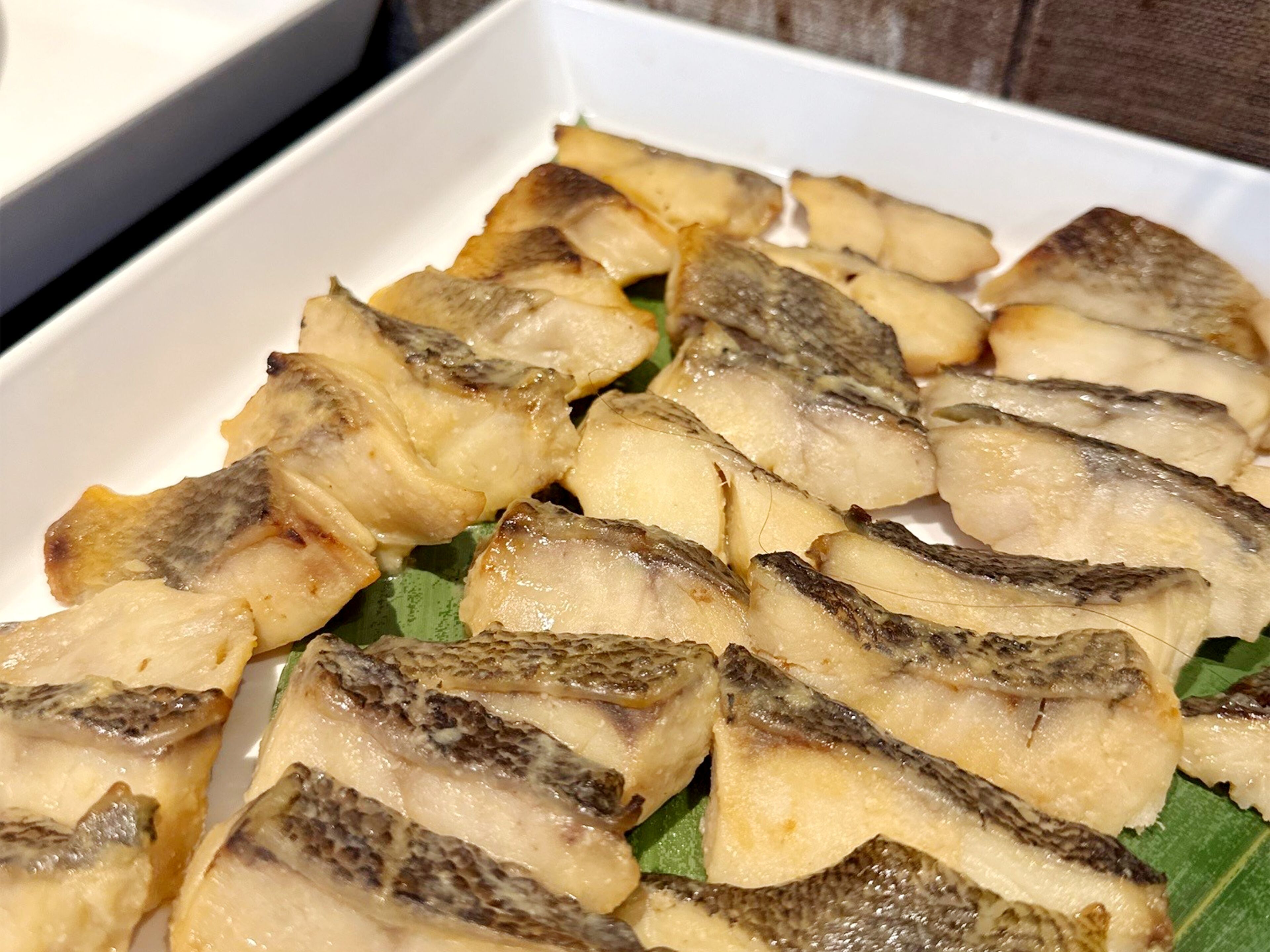 Une assiette rectangulaire blanche sur laquelle sont disposés plusieurs morceaux de poisson grillé, le tout posé sur une feuille verte.