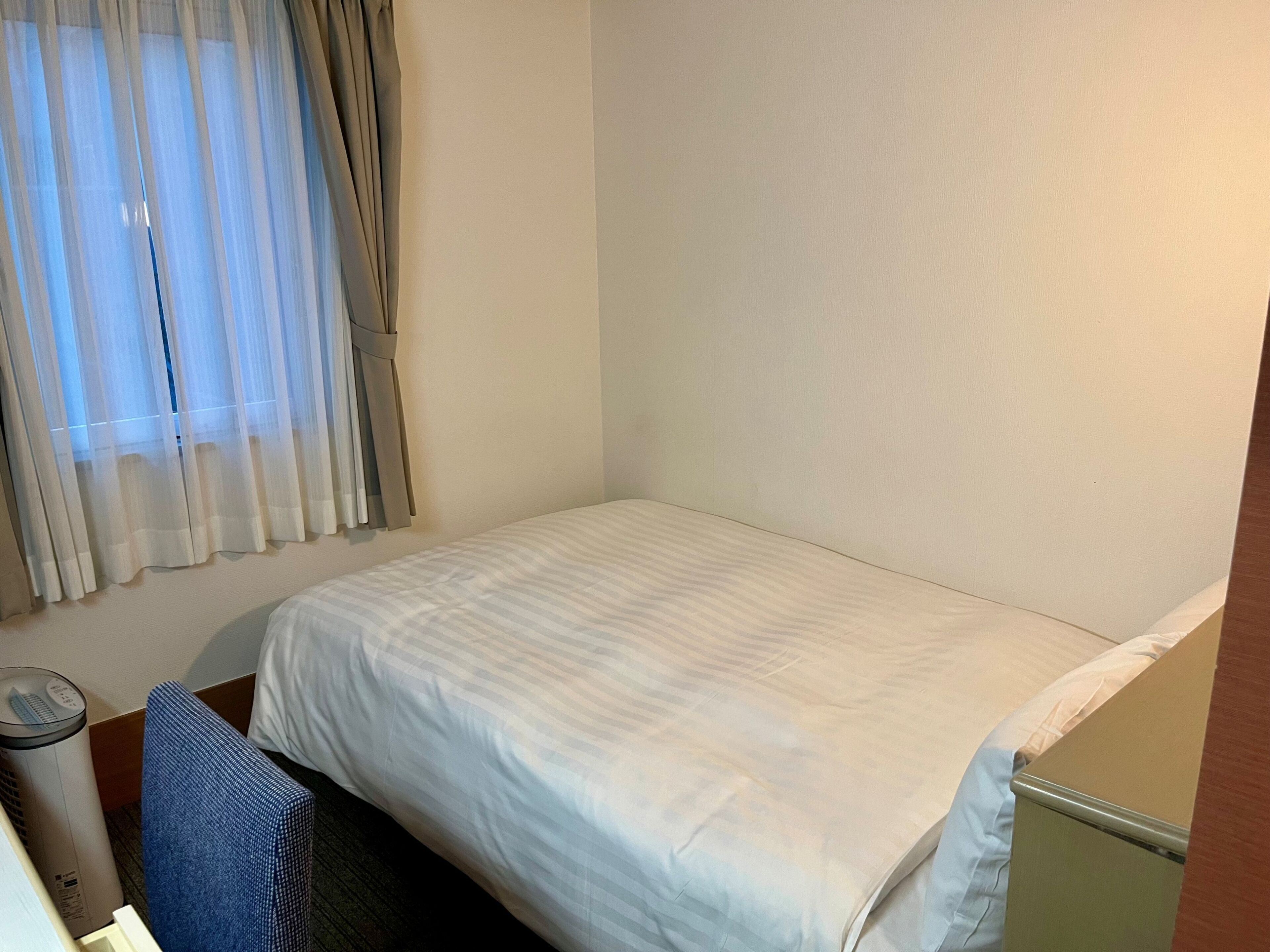 Une petite chambre avec un lit simple, un mur blanc uni, une fenêtre avec des rideaux légers, une table de chevet avec une lampe et une chaise bleue.