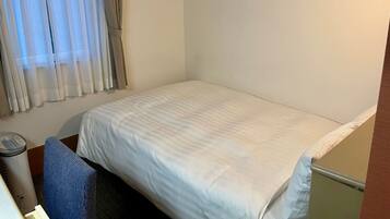 Une petite chambre avec un lit simple, un mur blanc uni, une fenêtre avec des rideaux légers, une table de chevet avec une lampe et une chaise bleue.