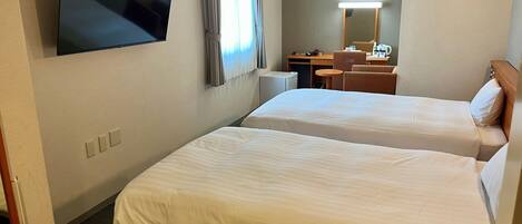 A hotel room with two beds, a TV mounted on the wall, a desk with a chair, and a small table with a lamp.