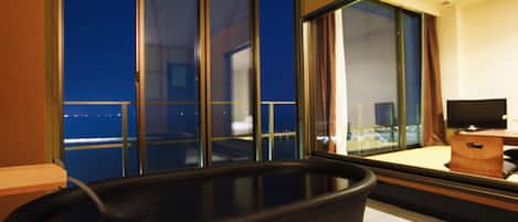 A room with a large window, a bathtub, a television, and a wooden bench.