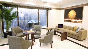 A modern hotel lobby with a sofa, chairs, a coffee table, and a large window offering a city view.