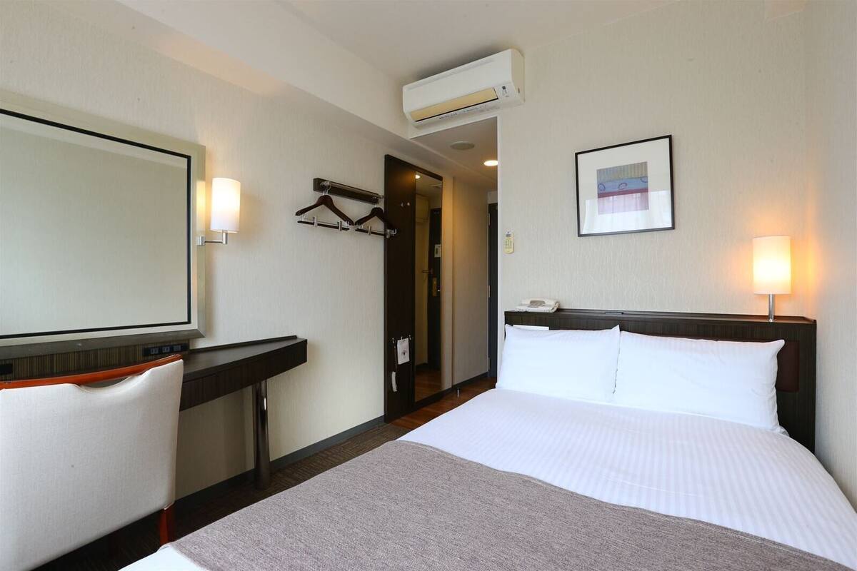 A hotel room with a bed, a desk, a chair, a mirror, a picture on the wall, and an air conditioning unit.
