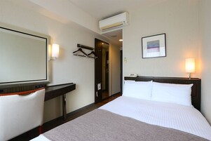 A hotel room with a bed, a desk, a chair, a mirror, a picture on the wall, and an air conditioning unit.