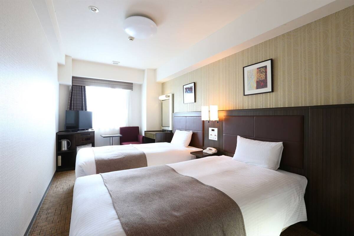 A hotel room with two beds, a desk, a chair, a television, and a window.