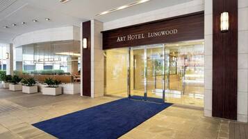 La entrada del Art Hotel Lungwood, con alfombra azul, puertas de cristal y un interior moderno.
