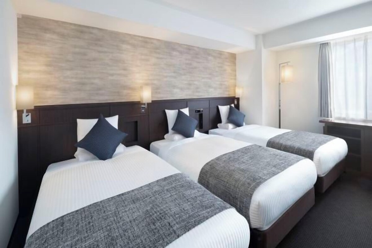 Habitación de hotel con tres camas, cada una con cabecero oscuro y ropa de cama blanca. Hay lámparas de pared a cada lado de las camas, y una ventana con cortinas a la derecha.