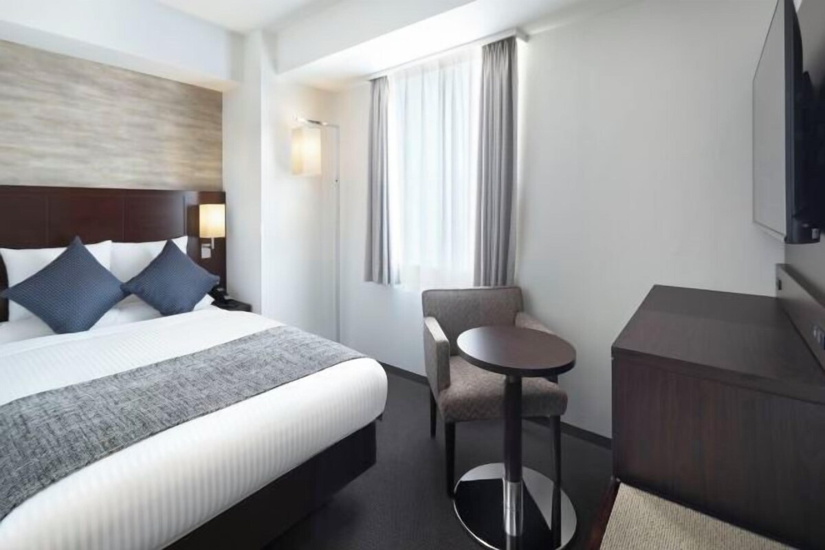Habitación de hotel con una cama grande, una mesita redonda pequeña, una silla, un televisor y una ventana con cortinas.