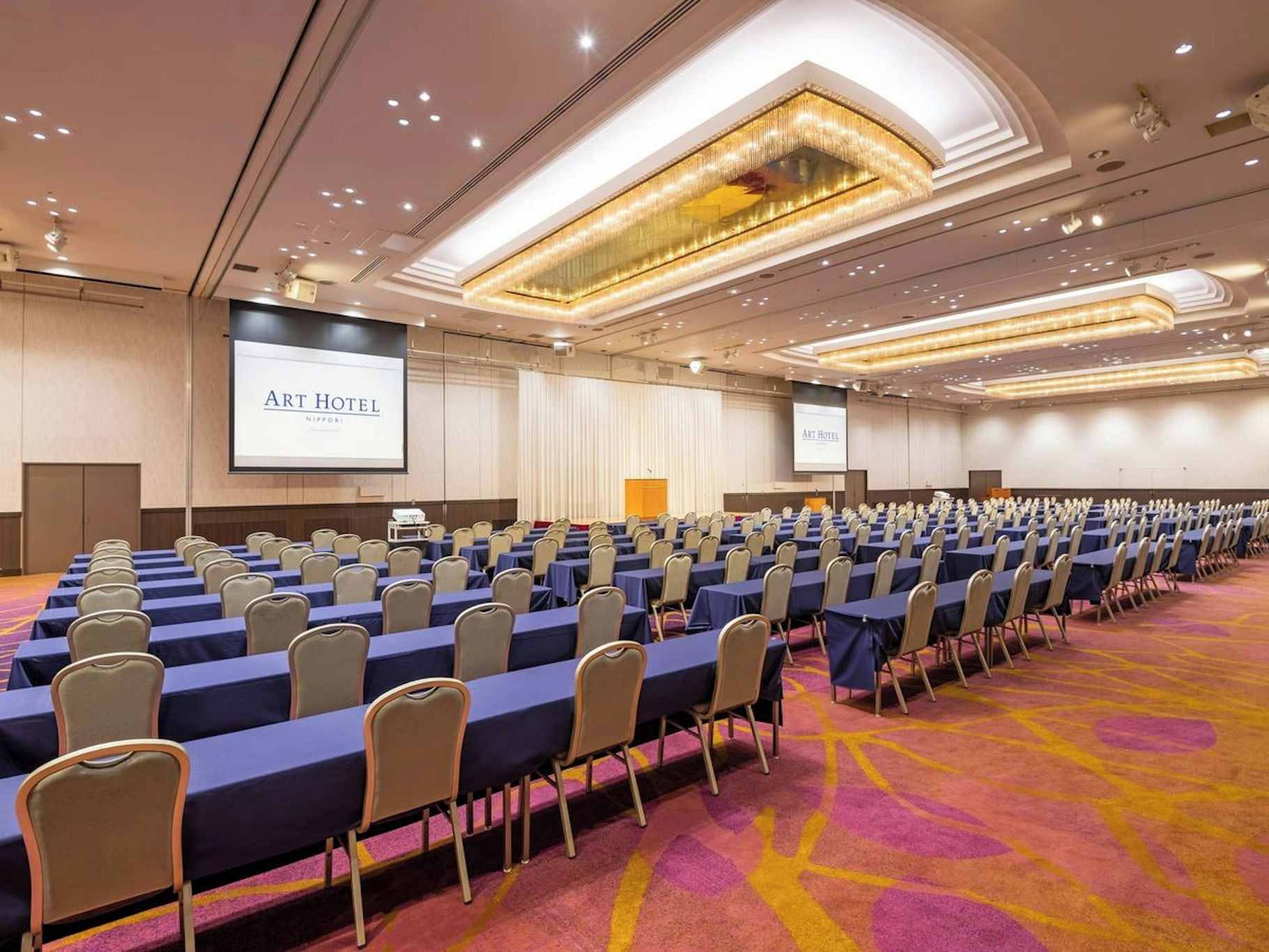 Un salón de conferencias con filas de mesas y sillas, una pantalla grande mostrando 'Art Hotel' y suelo alfombrado.