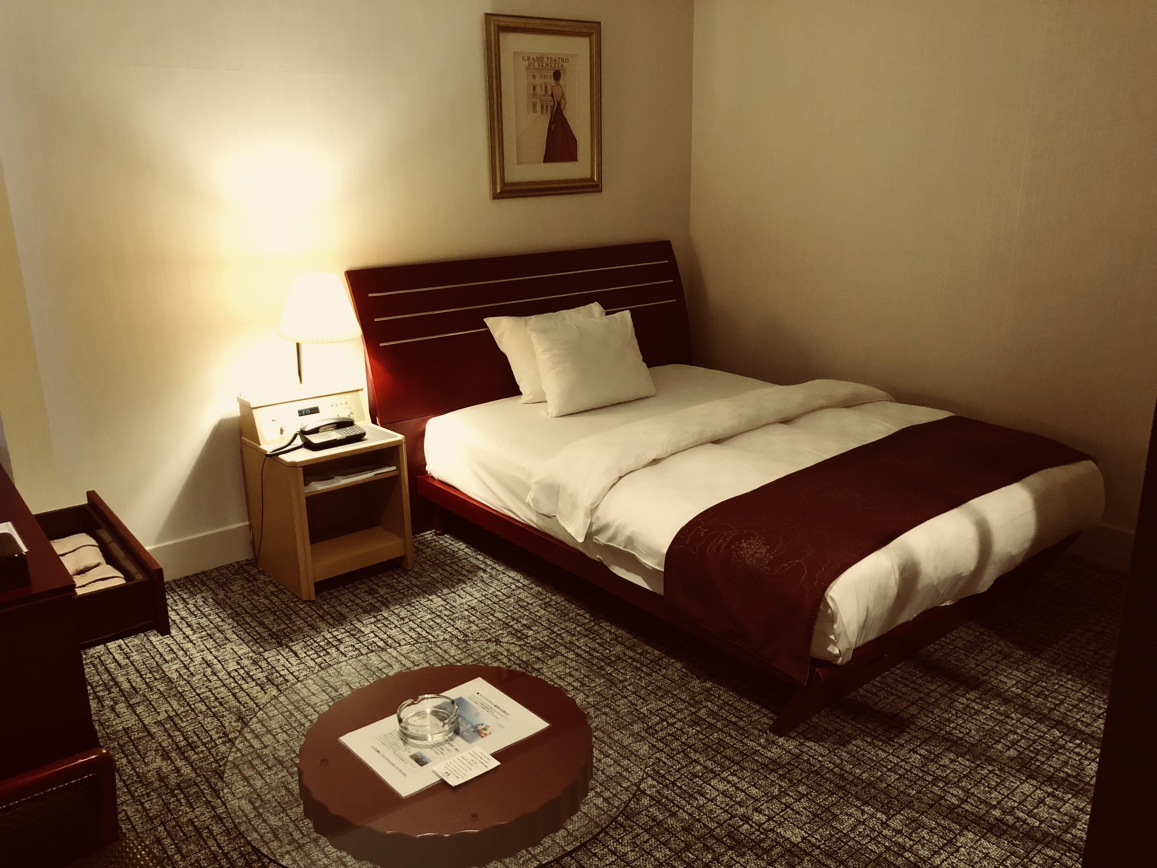 A hotel room with a single bed, a bedside table with a lamp, a small table with a menu, and a framed picture on the wall.