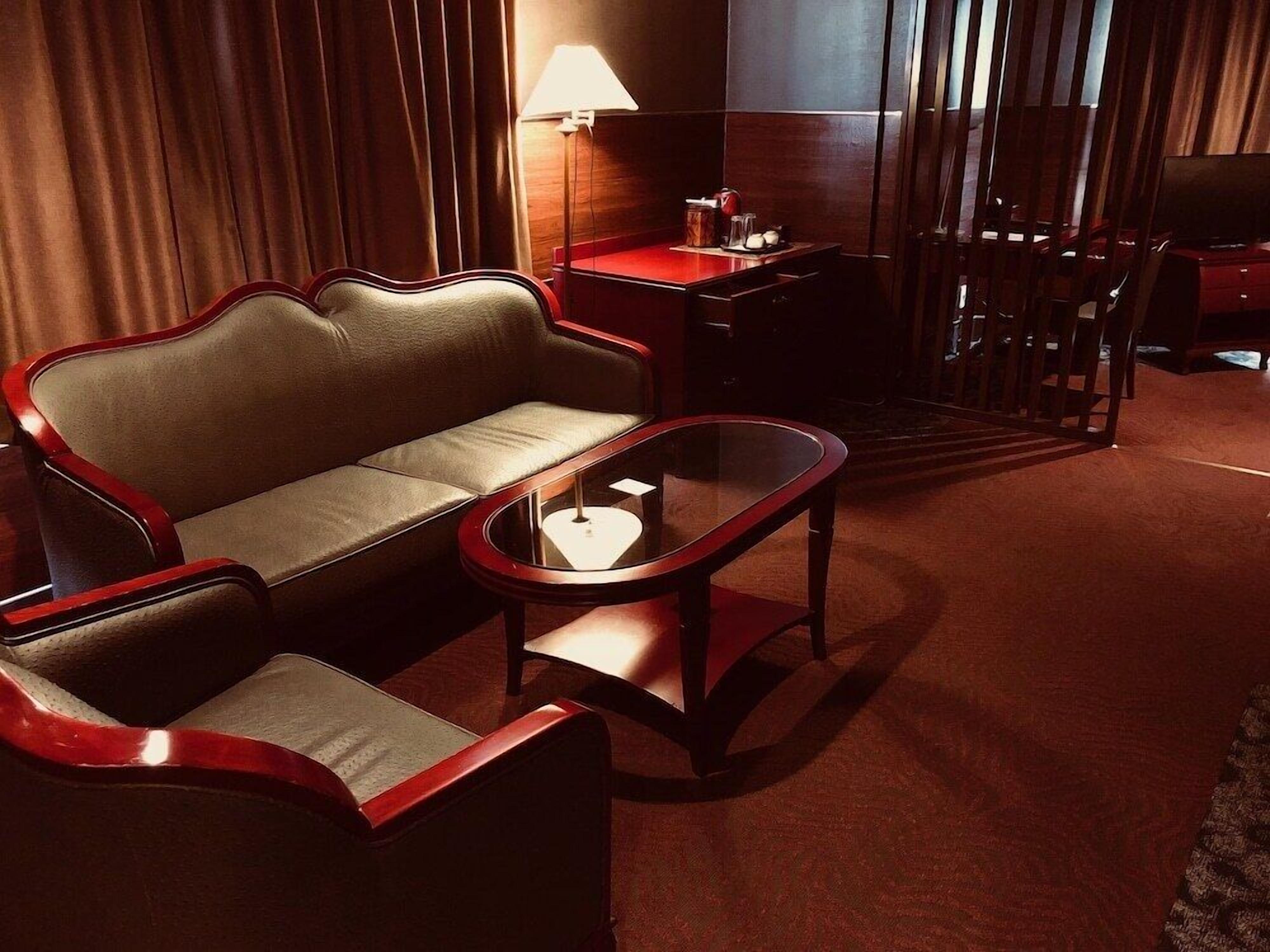 A hotel room with a leather sofa, a glass coffee table, a desk with a lamp, and a TV.