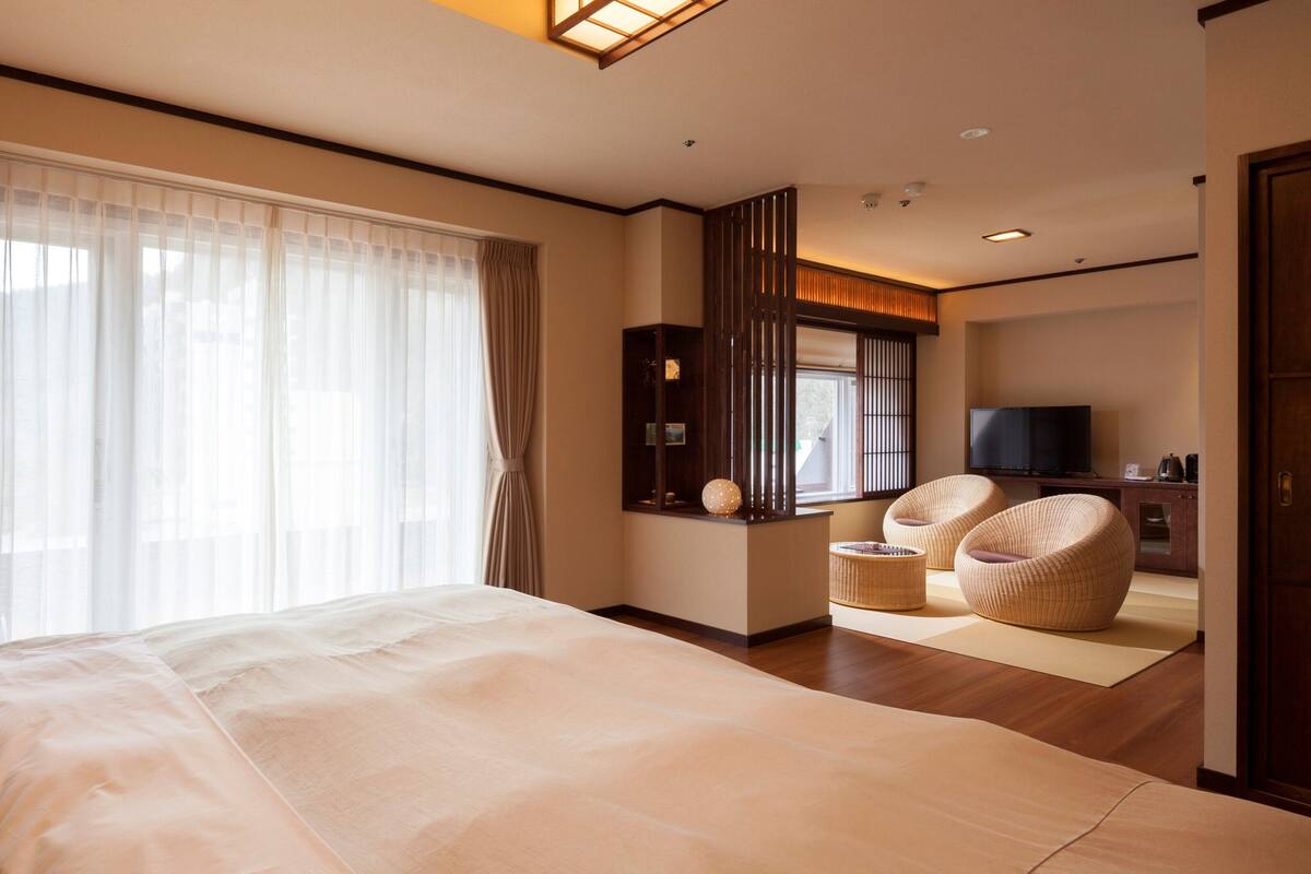 A hotel room with a large bed, a television, a seating area with chairs, and a view through the window.